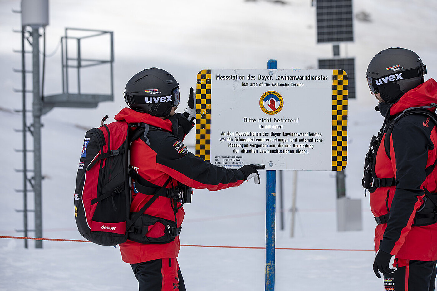 Zwei Skiwachtler stellen in einem Skigebiet ein Schild auf, das auf eine Messstation des Bayerischen Lawinenwarndienstes hinweist